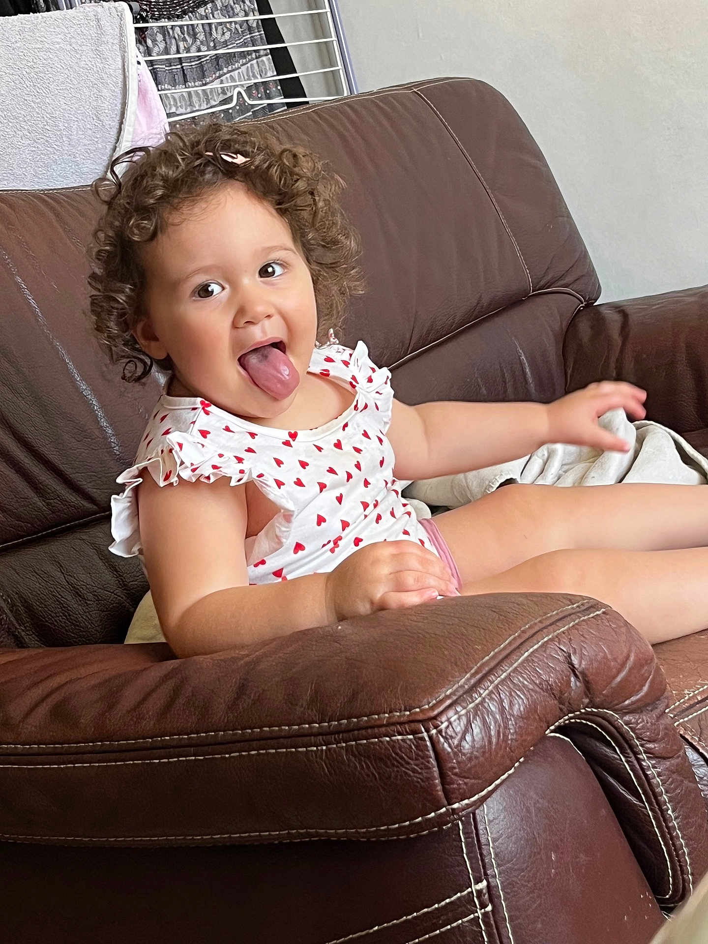 Juliette participe au concours pour gagner de l'argent avec cette photo : toddler, child, curly_hair, tongue_out, white_dress, red_hearts, brown_couch, leather, indoor, playful, smiling, person, furniture, home, relaxed, seated, clothing, face, expression, cute