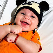 Ian is registered to the contest to win money with this photo: baby, smiling, infant, hat, mickey_mouse, orange_shirt, bracelet, happy, lying_down, white_background, measuring_scale, cute, child, portrait, close_up, clothing, headwear, person, baby_girl, baby_boy