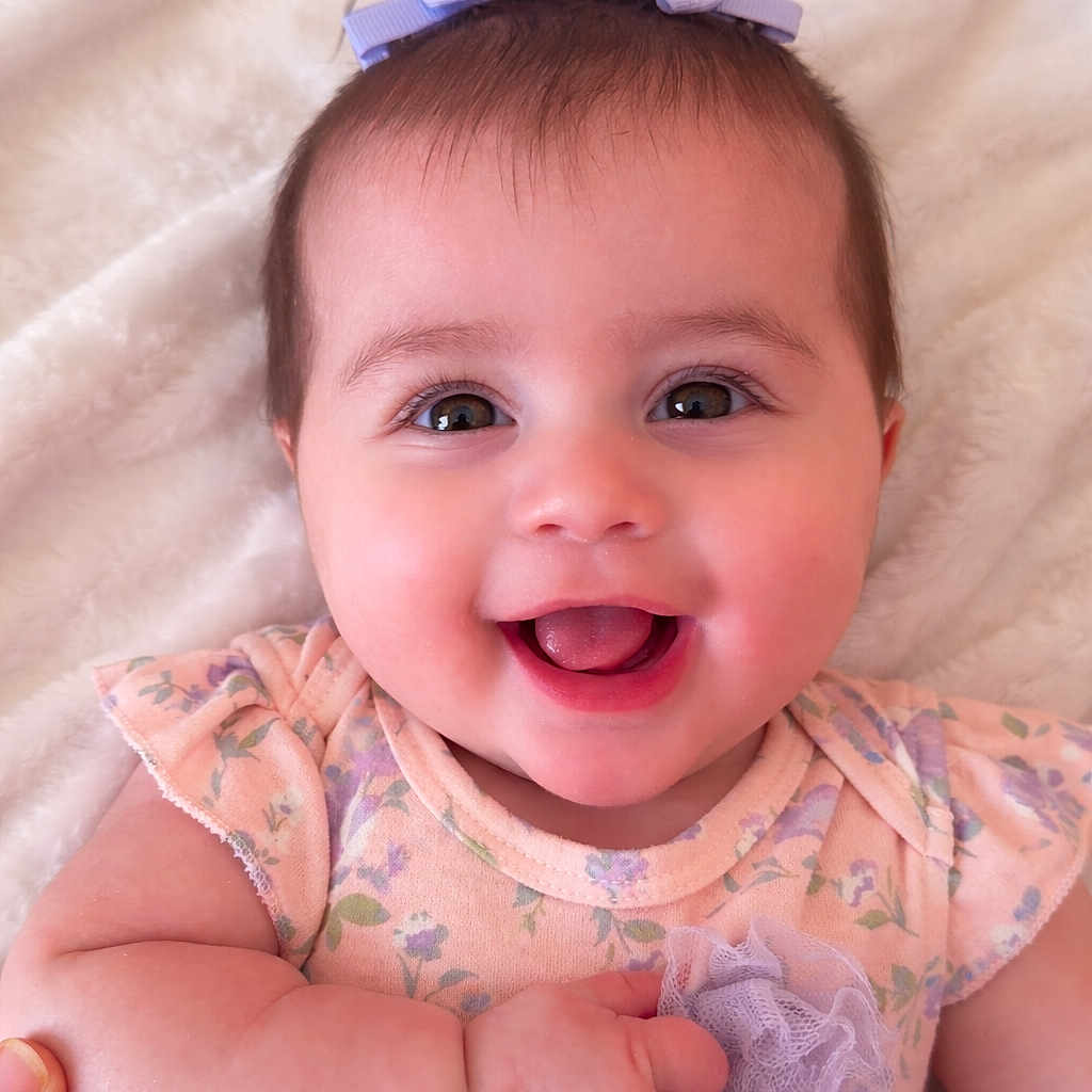 Melody is registered to the contest to win money with this photo: baby, child, smiling, face, headband, bow, flower, dress, floral, infant, cute, happy, soft_blanket, portrait, skin, hand, lying_down, indoors, closeup, person