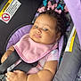 Lilly is registered to the contest to win money with this photo: baby, car_seat, pink_bib, curly_hair, earrings, child, infant, seatbelt, purple, black, warning_label, pacifier, cute, indoor, closeup, portrait, smile, clothing, skin, hand