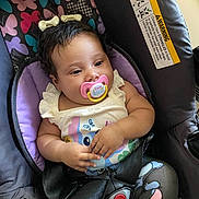 Lilly is registered to the contest to win money with this photo: baby, pacifier, car_seat, butterfly_pattern, child, infant, clothing, hair_bow, seatbelt, patterned_pants, cute, comfort, relaxed, indoors, person, young_child, sitting, closeup, portrait, colorful
