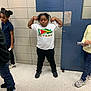 child, children, tshirt, flag, guyana, hallway, tile_wall, blue_door, floor, sneakers, black_pants, flexing, posing, casual, indoor, person, group, standing, young, school