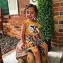 child, toddler, curly_hair, yellow_outfit, sunflower_pattern, cow_print, smiling, tortoise_statue, porch, brick_wall, green_shrub, barefoot, outdoor, happy, cute, summer_clothing, playful, decor, young_child, sitting