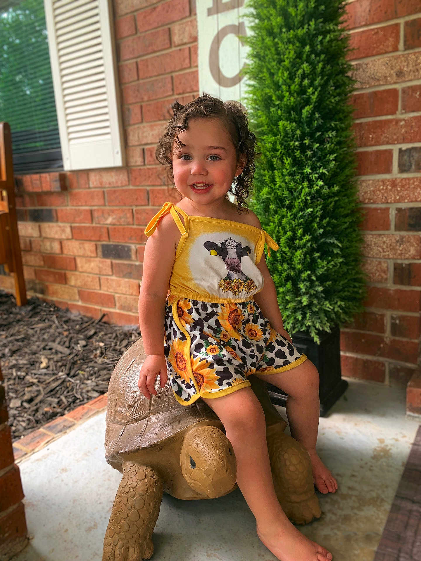 Eniyah is registered to the contest to win money with this photo: child, toddler, curly_hair, yellow_outfit, sunflower_pattern, cow_print, smiling, tortoise_statue, porch, brick_wall, green_shrub, barefoot, outdoor, happy, cute, summer_clothing, playful, decor, young_child, sitting