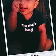 Zyair is registered to the contest to win money with this photo: baby, child, clothing, cute, dark_background, face, handwritten_date, happy, indoors, infant, mamas_boy, person, polaroid_frame, portrait, seated, smiling, text, thumbsucking, tshirt, young