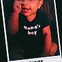 baby, child, clothing, cute, dark_background, face, handwritten_date, happy, indoors, infant, mamas_boy, person, polaroid_frame, portrait, seated, smiling, text, thumbsucking, tshirt, young