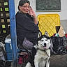 dog, husky, siberian_husky, person, woman, portrait, outdoor, sitting, leash, collar, tongue_out, fluffy, black_and_white, glasses, smiling, chalkboard, cooler, plastic_chair, backyard, pets