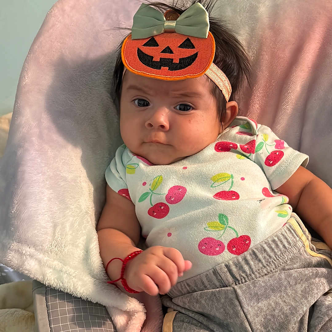 Keitlyn Ramírez joined the competition — help win amazing prizes! baby, cherry_pattern, child, clothing, comfortable, cozy, cute, expression, headwear, indoor, infant, pants, person, portrait, pumpkin_headband, red_bracelet, seated, shirt, soft_cushion, young