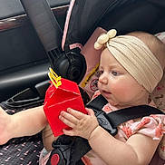Raya is registered to the contest to win money with this photo: baby, infant, child, car_seat, headband, bow, happy_meal_box, red_box, car_interior, seatbelt, pink_clothing, floral_pattern, hand, foot, curious, portrait, sitting, safety_harness, cute, young_child