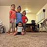 children, siblings, baby, toddler, family, group_portrait, indoor, living_room, sitting, standing, smiling, portrait, casual_clothing, red_shirt, blue_shirt, pink_shirt, carpet, baby_equipment, window_blinds, home