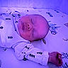 baby, infant, sleeping, lying_down, face, portrait, closed_eyes, onesie, clothing, blanket, bedding, bed, indoors, purple_lighting, patterned_fabric, hands, peaceful, cute, resting, photography