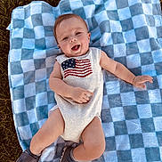 Hudson is registered to the contest to win money with this photo: baby, infant, blanket, checkered, outdoor, grass, smile, happy, onesie, american_flag, shoe, footwear, lying_down, child, portrait, cute, young, person, summer, sunlight