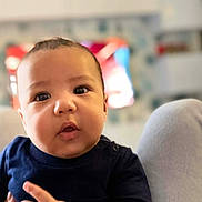 Jayden a rejoint le concours — aidez-le/la à gagner de superbes lots ! baby, blurred_background, bokeh, cheeks, child, cute, expression, eyes, face, hand, indoors, infant, living_room, mouth, navy_shirt, onesie, portrait, skin, soft_lighting, television
