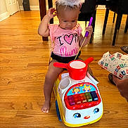 Gianna is registered to the contest to win money with this photo: toddler, child, indoor, toy_car, party_hat, drum, purple_drumstick, pink_shirt, wooden_floor, furniture, chair, floor, playtime, cute, baby, smiling, person, celebration, colorful_toy, happy