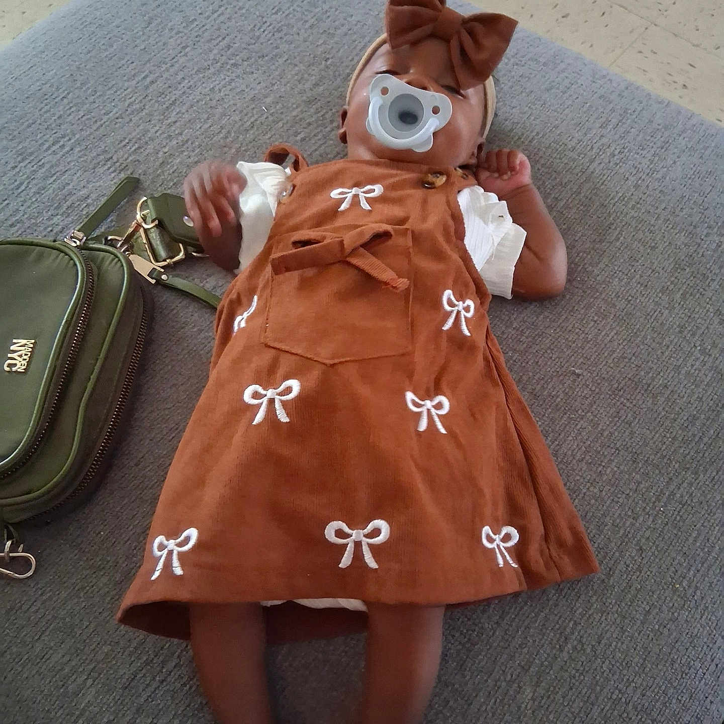 Zia joined the competition — help win amazing prizes! baby, infant, child, pacifier, bow, headband, dress, brown_clothing, white_sleeves, lying_down, couch, handbag, fashion, portrait, indoor, carpet, furniture, feet, baby_doll, photography