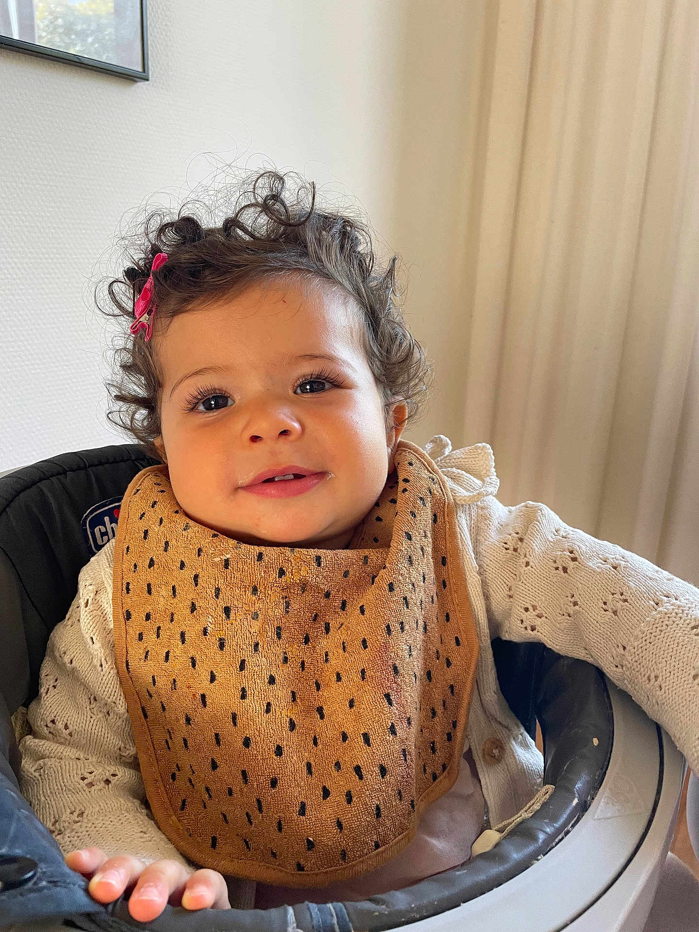 Inaya a rejoint le concours — aidez-le/la à gagner de superbes lots ! toddler, child, curly_hair, pink_hair_clip, bib, knitted_sweater, high_chair, smile, face, hand, indoors, window, curtain, baby, person, clothing, seated, happy, home, portrait