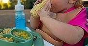 Winter is registered to the contest to win money with this photo: child, toddler, baby, eating, corn, food, high_chair, bib, pink_shirt, outdoor, picnic, plate, macaroni, vegetables, drink, bottle, smiling, portrait, summer, family_meal