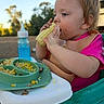 child, toddler, baby, eating, corn, food, high_chair, bib, pink_shirt, outdoor, picnic, plate, macaroni, vegetables, drink, bottle, smiling, portrait, summer, family_meal