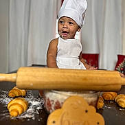 Keyler is registered to the contest to win money with this photo: baby, child, chef_hat, apron, croissant, flour, rolling_pin, baking, table, kitchen, smiling, face, food, wooden_sign, celebration, portrait, indoor, cute, happy, person