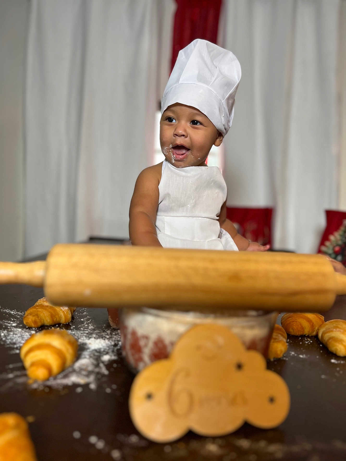 Keyler is registered to the contest to win money with this photo: baby, child, chef_hat, apron, croissant, flour, rolling_pin, baking, table, kitchen, smiling, face, food, wooden_sign, celebration, portrait, indoor, cute, happy, person