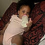 baby, bottle, curly_hair, onesie, pillow, lying_down, indoor, child, infant, drink, patterned_textile, pink, hand, face, sleepy, cozy, relaxed, clothing, cute, person