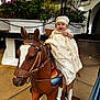 Milton joined the competition — help win amazing prizes! baby, child, smiling, wooden_horse, statue, outdoor, plants, greenery, white_building, dress, hat, happy, person, sitting, playful, decorative, floor, daytime, cute, portrait