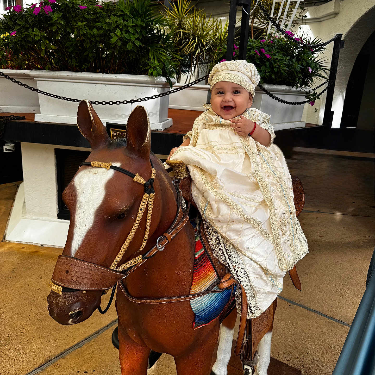 Milton joined the competition — help win amazing prizes! baby, child, cute, daytime, decorative, dress, floor, greenery, happy, hat, outdoor, person, plants, playful, portrait, sitting, smiling, statue, white_building, wooden_horse