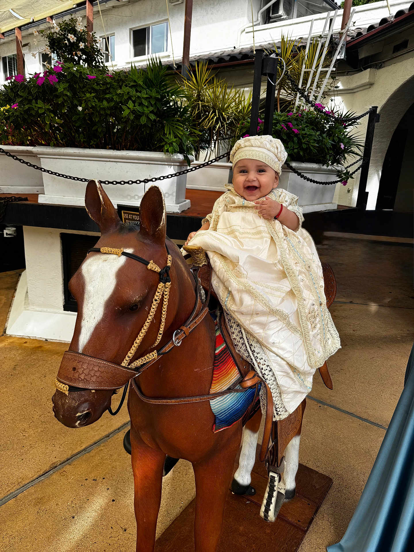 Milton joined the competition — help win amazing prizes! baby, child, smiling, wooden_horse, statue, outdoor, plants, greenery, white_building, dress, hat, happy, person, sitting, playful, decorative, floor, daytime, cute, portrait