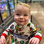 baby, blurry_background, child, clothing, cute, expression, face, hand, holiday_theme, indoor, infant, pajamas, person, retail, seatbelt, shopping_cart, smiling, store, supermarket, young