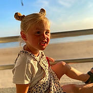 Lya participe au concours pour gagner de l'argent avec cette photo : child, girl, blonde_hair, double_buns, smiling, sunlight, beach, sea, sand, summer, casual_clothing, white_tshirt, floral_overall, outdoor, happy, portrait, sitting, daylight, nature, relaxation