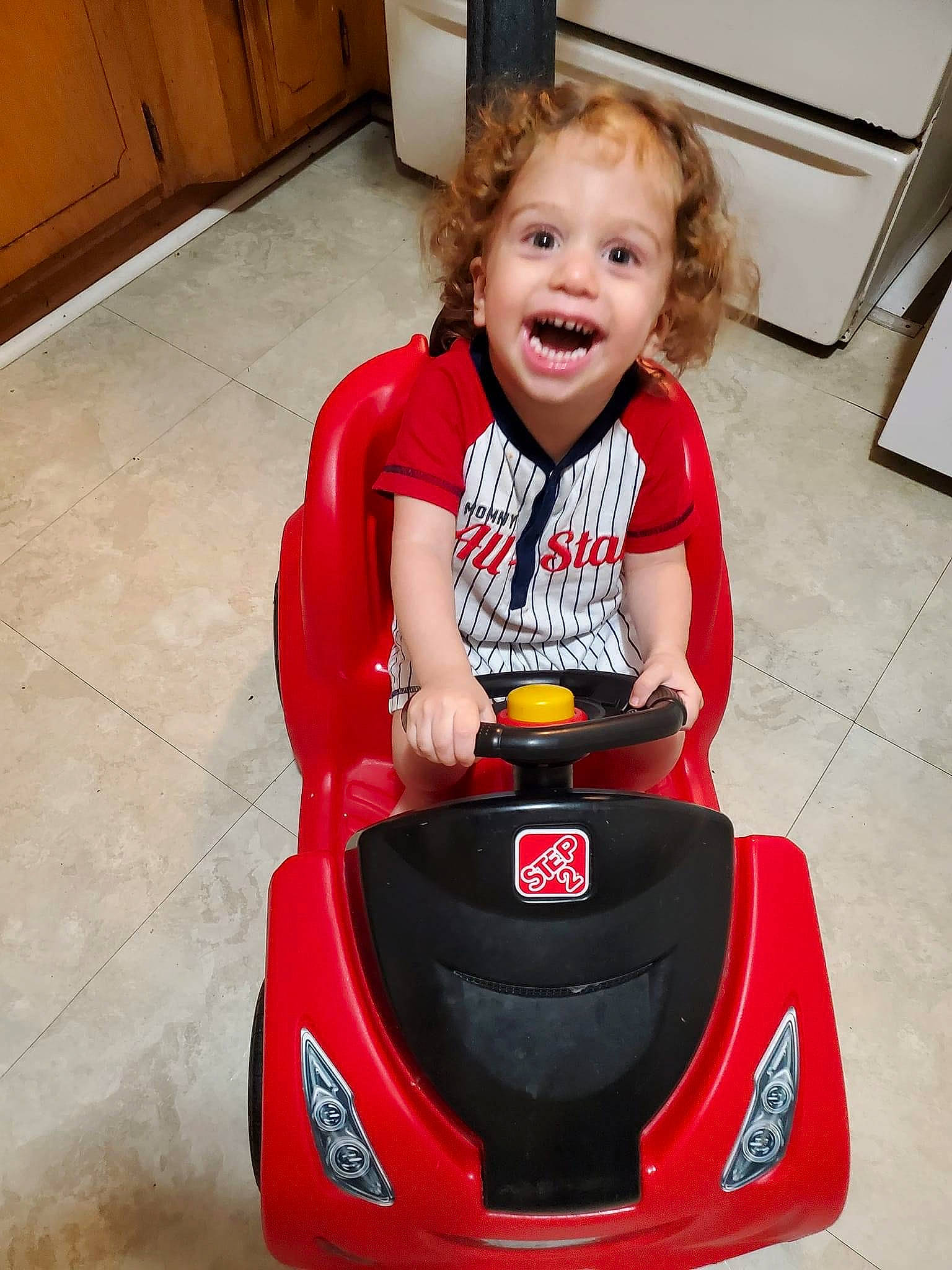 Gabriel is registered to the contest to win money with this photo: automotive_design, automotive_exterior, automotive_tire, bumper, cabinetry, face, floor, flooring, fun, happy, hardwood, motor_vehicle, person, riding_toy, smile, tire, toddler, vehicle, white, wood