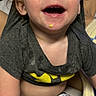 toddler, child, smiling, happy, batman_shirt, messy_face, food_on_chin, short_sleeves, pants, indoor, blanket, seated, person, cute, young_child, casual_clothing, light_skin, hair, cheerful, portrait