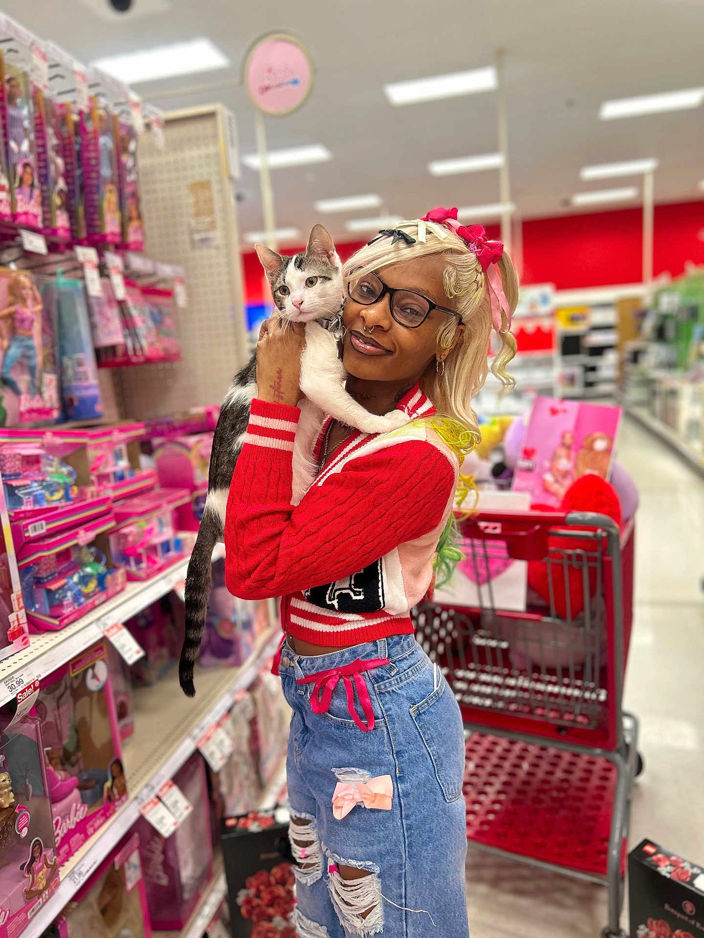 Stormei is registered to the contest to win money with this photo: person, cat, shopping_cart, toy_aisle, dolls, retail_store, blonde_hair, glasses, septum_piercing, colorful_clothing, red_sweater, ripped_jeans, bow, pink, holding_pet, portrait, smiling, aisle, shelves, playful