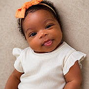 Andrea Rene joined the competition — help win amazing prizes! baby, infant, smile, tongue_out, headband, orange_bow, white_clothing, ruffled_sleeves, curly_hair, portrait, closeup, cute, happy, child, face, skin, eyes, soft_background, lying_down, adorable