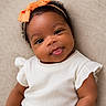 baby, infant, smile, tongue_out, headband, orange_bow, white_clothing, ruffled_sleeves, curly_hair, portrait, closeup, cute, happy, child, face, skin, eyes, soft_background, lying_down, adorable