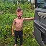 boy, child, barefoot, shirtless, mohawk, grass, yellow_flowers, car_door, outdoor, nature, greenery, pants, smiling, young, person, daylight, recreational_vehicle, casual, standing, happy