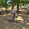 child, boy, girl, statue, deer_statue, park, trees, shade, sunlight, fence, picnic_table, sand, outdoor, playground, casual_clothing, smiling, daytime, nature, recreation, fun