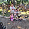 family, children, woman, dinosaur_statues, outdoor, park, tree, sunlight, grass, stroller, smiling, casual_clothing, footwear, barefoot, fence, nature, daytime, tattoo, glasses, posing