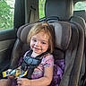 Hazel is registered to the contest to win money with this photo: child, car_seat, girl, purple_dress, snack, car_interior, seatbelt, window, greenery, smile, messy_hair, toddler, happy, sitting, daylight, face, clothing, person, seat, safety