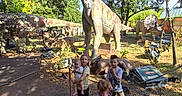 Alyssa joined the competition — help win amazing prizes! children, baby, dinosaur_statues, outdoor, trees, daylight, park, playground, kids, nature, blue_sky, shadows, grass, rocks, walking_path, summer_clothing, family, curious, greenery, sculpture