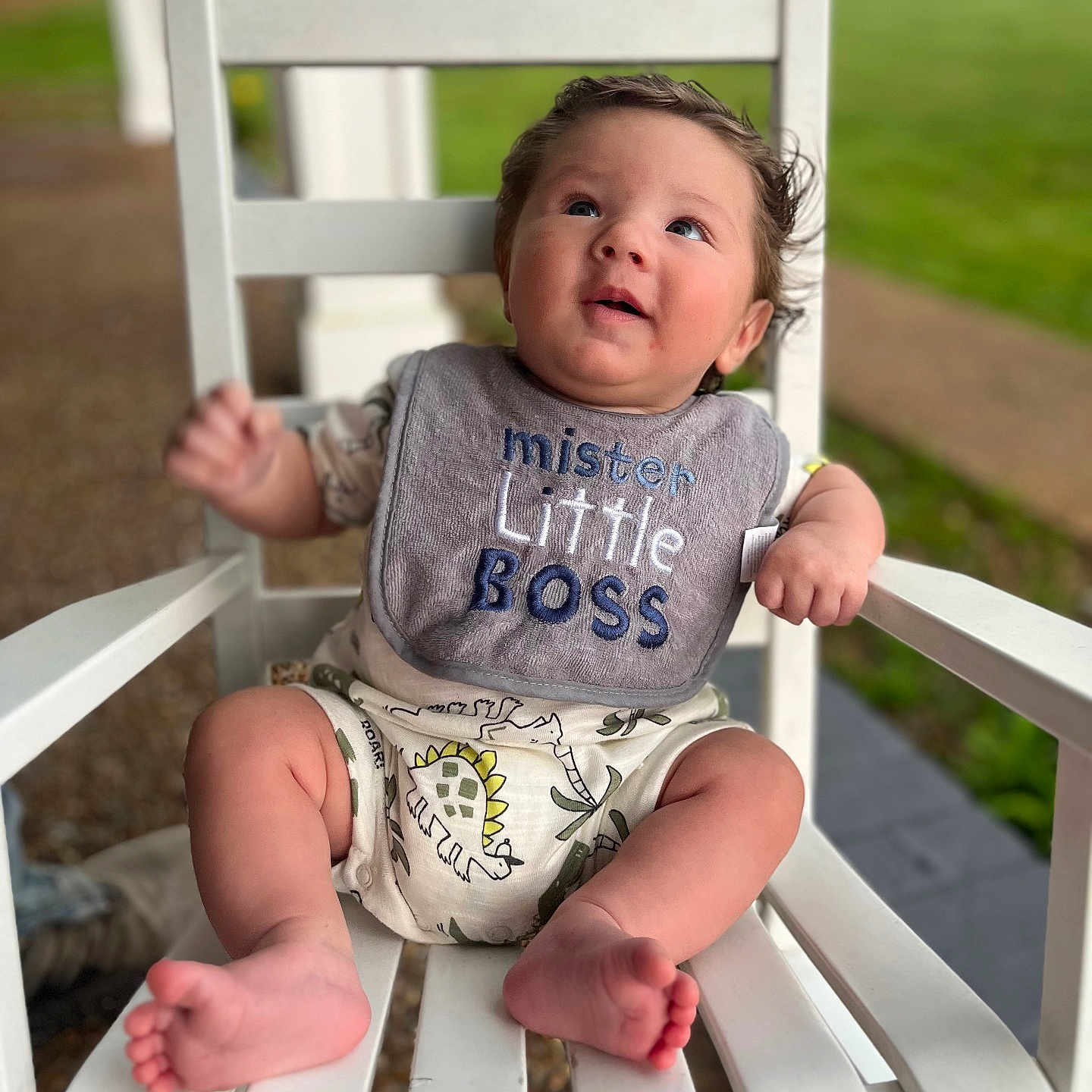 Christian-Wayne is registered to the contest to win money with this photo: baby, bib, child, clothing, cute, daylight, dinosaur_print, expression, face, feet, grass, hands, infant, outdoor, person, portrait, sitting, summer, white_chair, wooden_chair