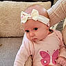 adult_hand, baby, bow, butterfly_print, child, couch, cozy, curious_expression, cushion, cute, hand, headband, indoors, infant, person, pink_clothing, portrait, seated, skin, soft_fabric
