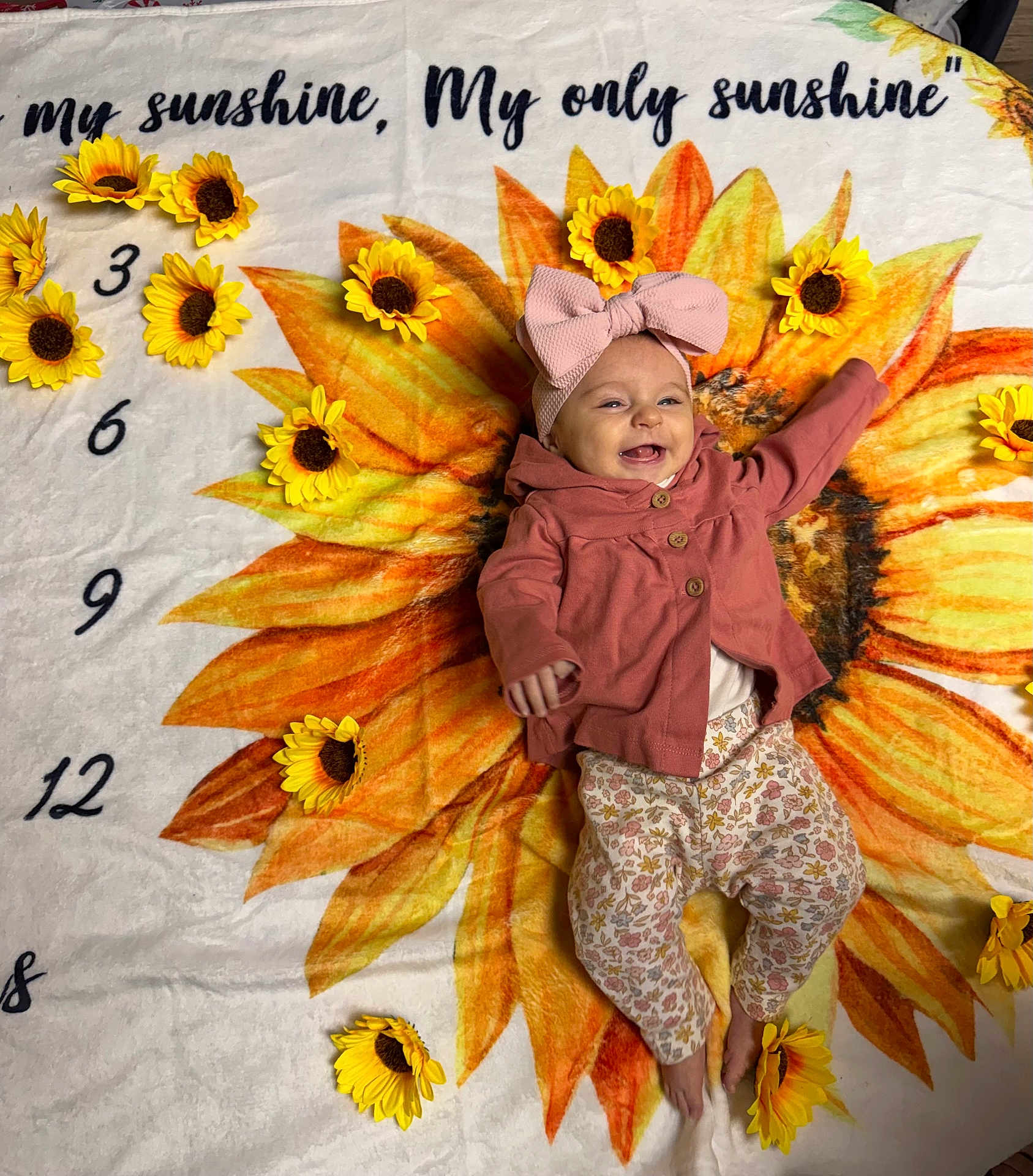 Athena joined the competition — help win amazing prizes! baby, child, smiling, sunflower, blanket, flower, bow, pink, clothing, floral, pattern, happy, infant, cute, lying, decor, yellow, orange, face, person