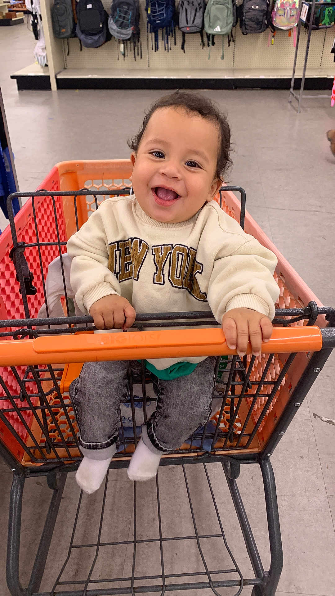 Erick is registered to the contest to win money with this photo: toddler, child, shopping_cart, smile, happy, sweatshirt, new_york, clothing, jeans, socks, store, backpacks, aisle, floor, handrail, person, indoor, cute, baby, retail