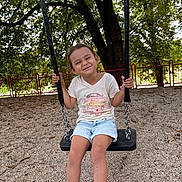 Arya participe au concours pour gagner de l'argent avec cette photo : child, swing, playground, smiling, happy, tree, gravel, fence, outdoor, summer, casual_clothing, shorts, sneakers, sock, person, nature, daylight, leisure, recreation, fun