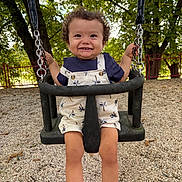 Nael a rejoint le concours — aidez-le/la à gagner de superbes lots ! toddler, child, swing, playground, smiling, curly_hair, overalls, shoes, socks, trees, fence, gravel, outdoor, daylight, happy, person, seat, metal_chain, nature, park
