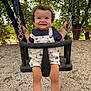 child, curly_hair, daylight, fence, gravel, happy, metal_chain, nature, outdoor, overalls, park, person, playground, seat, shoes, smiling, socks, swing, toddler, trees