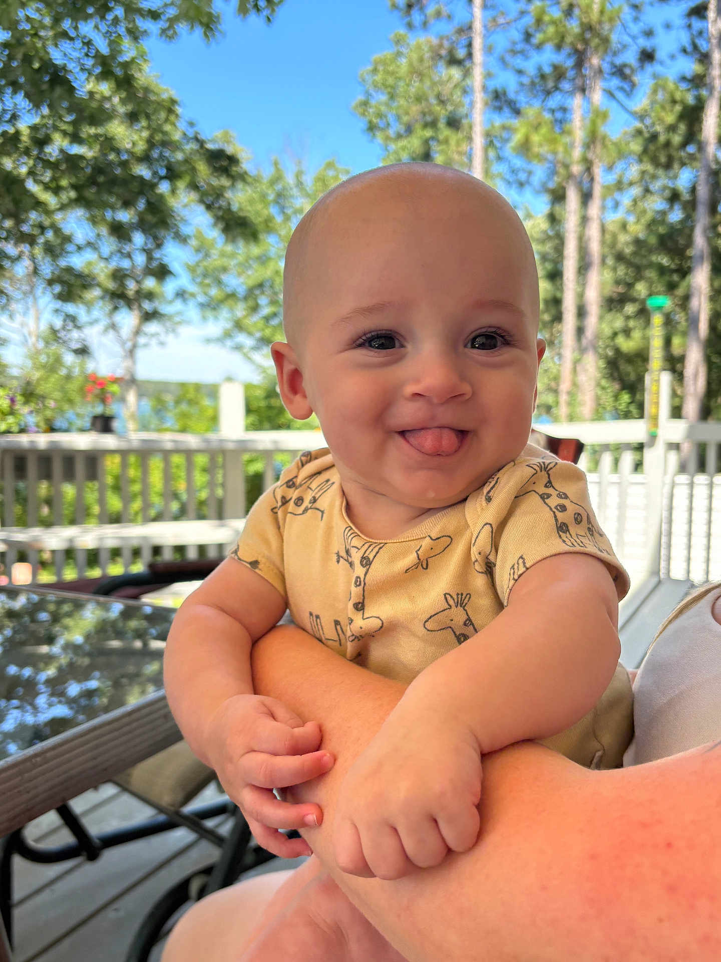 Fynn joined the competition — help win amazing prizes! baby, infant, child, smiling, tongue_out, outdoor, daylight, trees, deck, railing, person, holding, cute, happy, shirt, giraffe_print, skin, face, arm, nature