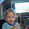 Cody is registered to the contest to win money with this photo: blue_shirt, box, cabinet, cartoon, child, clock, curious, entertainment, furniture, home, indoors, light_hair, living_room, person, playful, pointing, smartphone, smile, tongue, tv