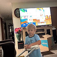 Cody is registered to the contest to win money with this photo: blue_shirt, blur, cabinet, cardboard_box, carpet, cartoon, child, clock, diaper, floor, flowers, furniture, home_interior, living_room, plastic_toy, television, toddler, toy_walker, vase, window